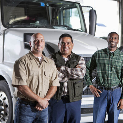 Three truck drivers standing in front of the cab of a white semi-truck parked in the garage of a warehouse.  The group of multracial men are standing in a row, smiling at the camera, looking relaxed and confident.  They are wearing blue jeans and button-down shirts.  The man in the middle is Hispanic.